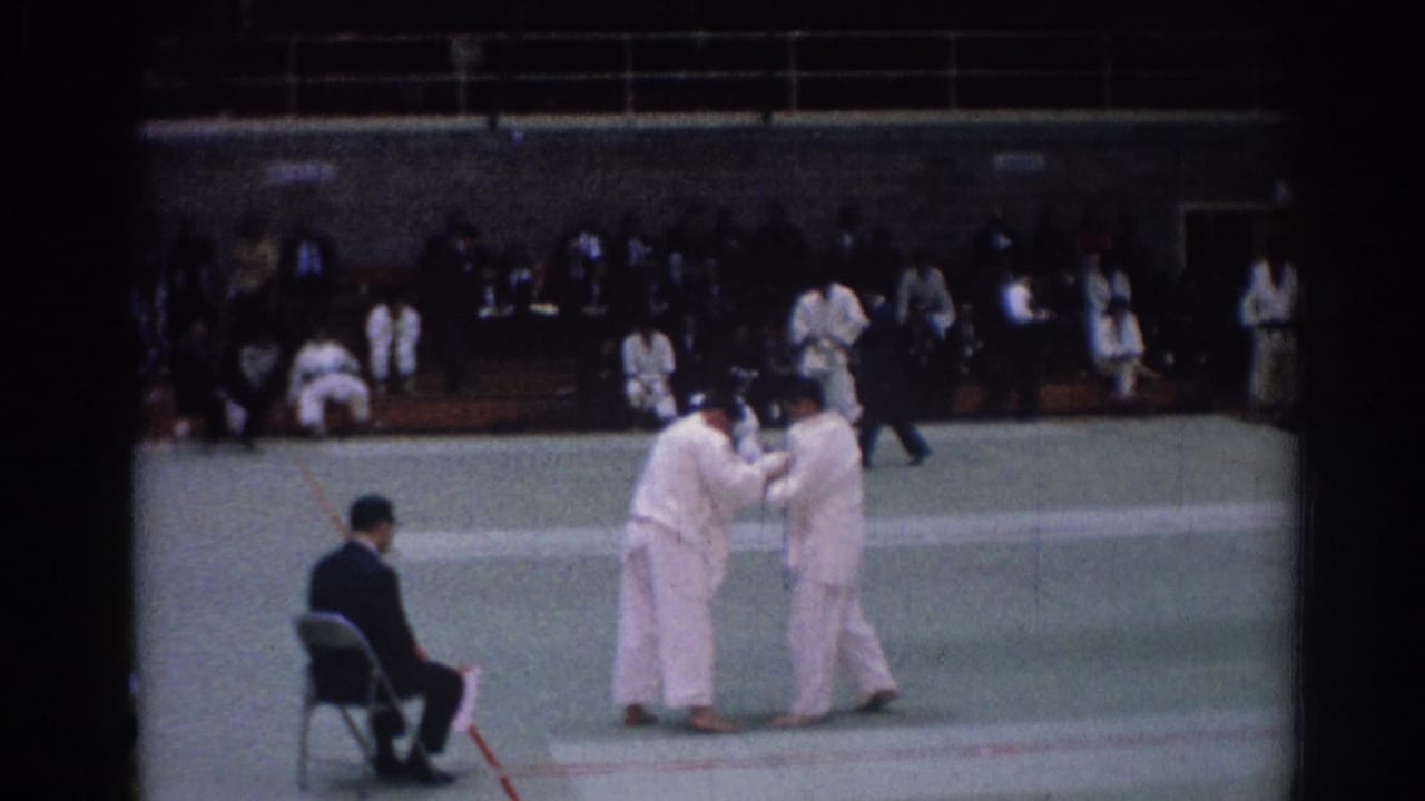 1967 Los Angeles judo match frame with paired athletes in white gi, a seated official, and spectators beyond the mat.