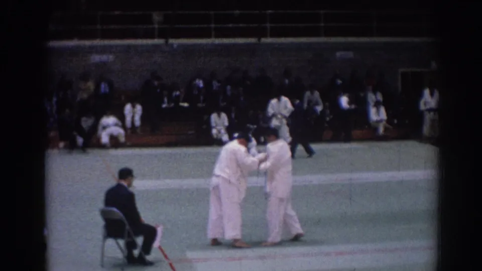 1967 Los Angeles judo match frame with paired athletes in white gi, a seated official, and spectators beyond the mat.