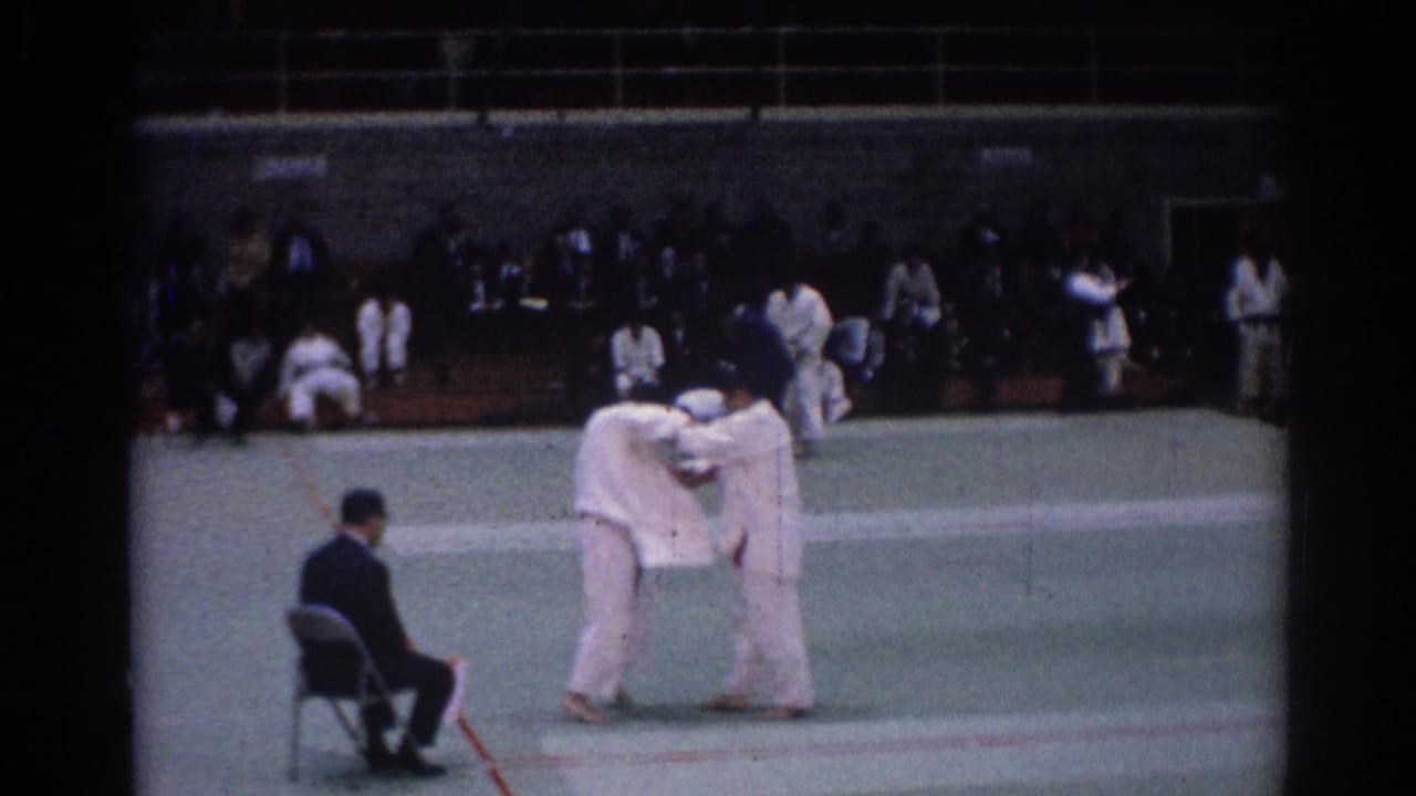 Two judo competitors grapple on a gym mat while a suited official sits nearby.
