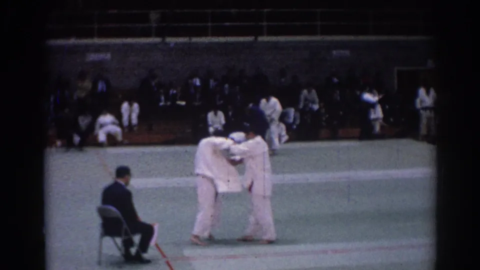 1967 Los Angeles home-movie frame of two judo competitors in white gi grappling on a green mat with a seated official nearby.