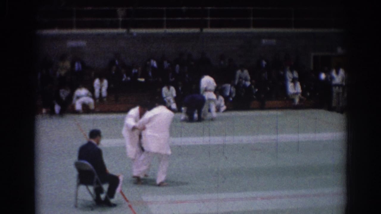 Judo competitors move across the mat with a seated official and spectators behind them.