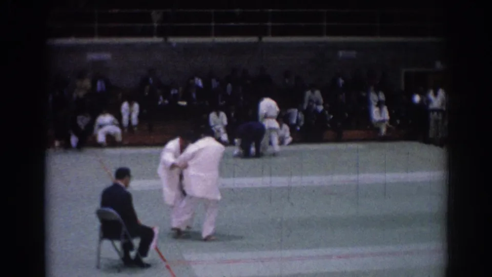 Two white-clad judo competitors shift position on the mat during a 1967 Los Angeles indoor match, with spectators in the background.