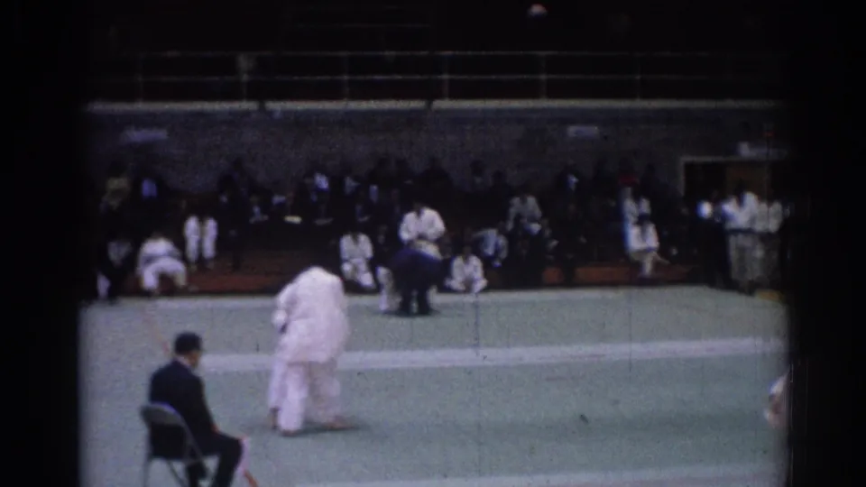 A 1967 indoor judo bout in Los Angeles continues as one competitor bends forward on the mat and spectators sit along the wall.
