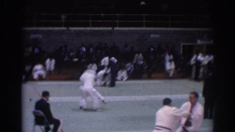 Late-frame view of the 1967 Los Angeles judo match, with one competitor moving on the mat and another white-clad participant at the right edge.