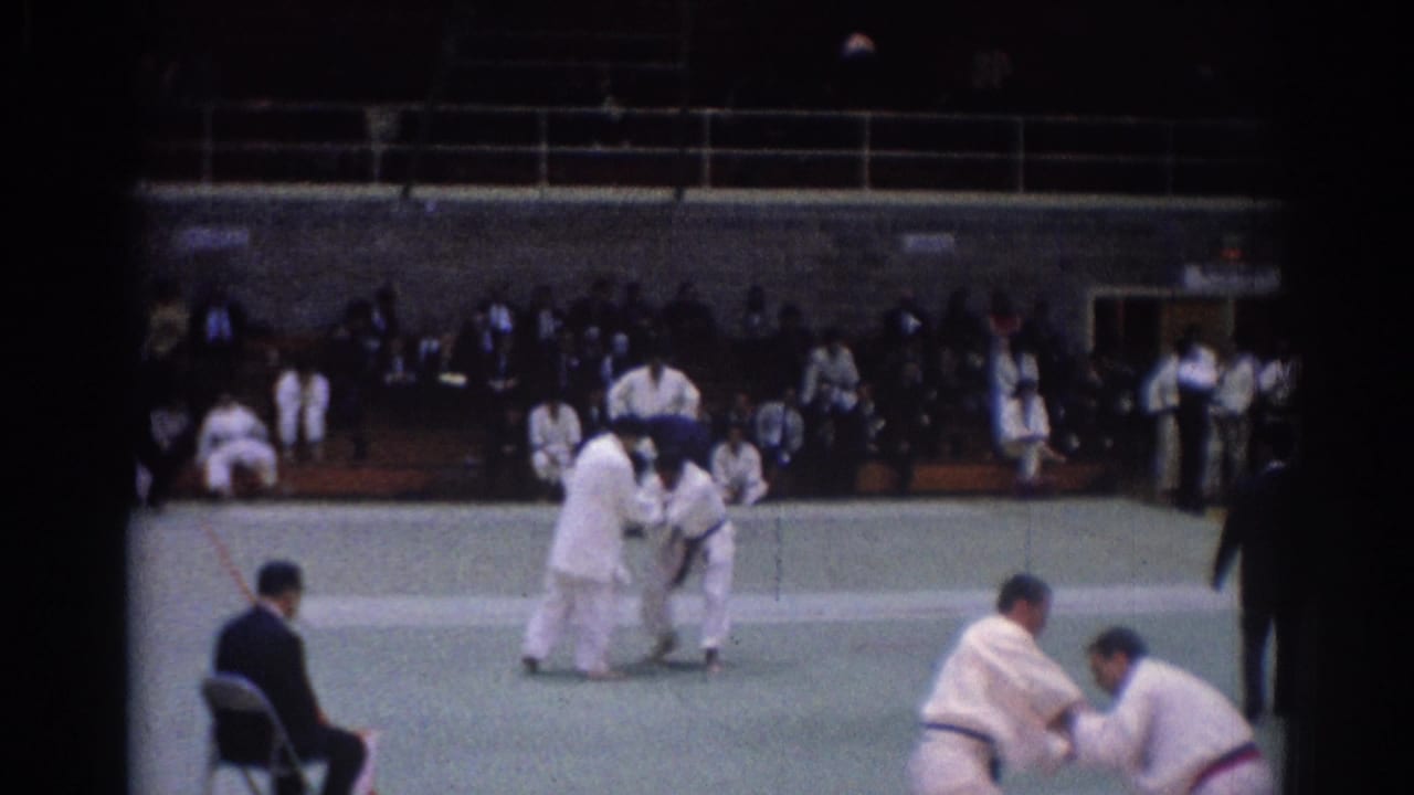 Judo competitors in white gi spar on a green mat as a referee and spectators watch.
