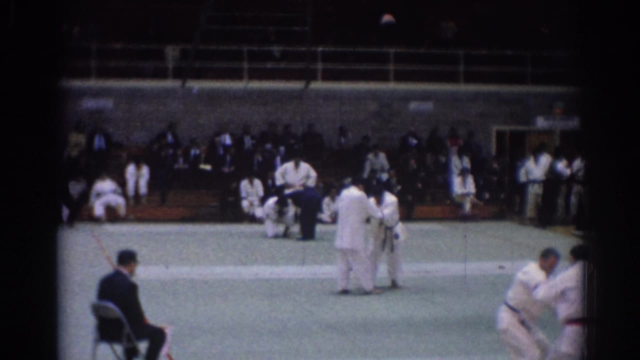 Two judo competitors stand on the mat while another pair grapples near the right edge.