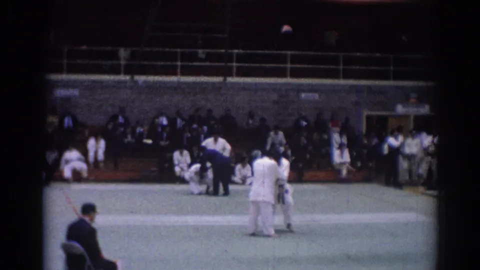 Referee attends to judo competitors during a 1967 Los Angeles indoor match, with spectators seated behind the green mat.