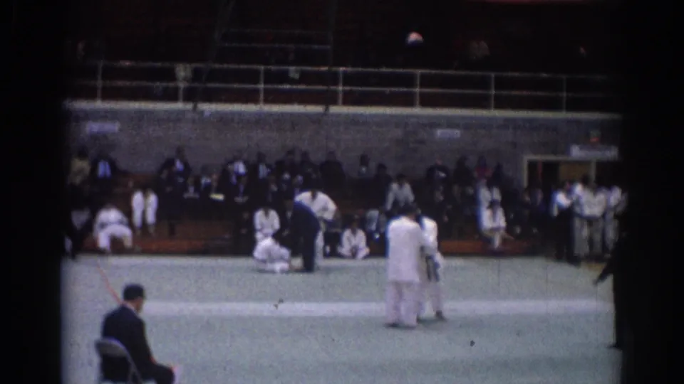 White-gi judo competitors stand during a 1967 Los Angeles indoor match, framed by a referee, green mat, and bleacher spectators.