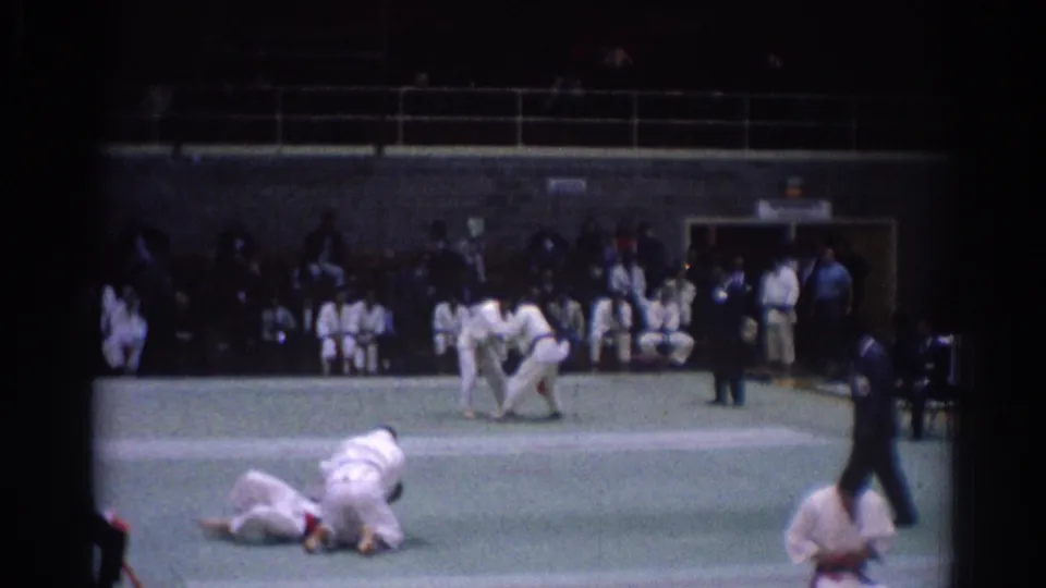 1967 Los Angeles judo competition view with athletes in white uniforms grappling on a green mat before seated spectators.