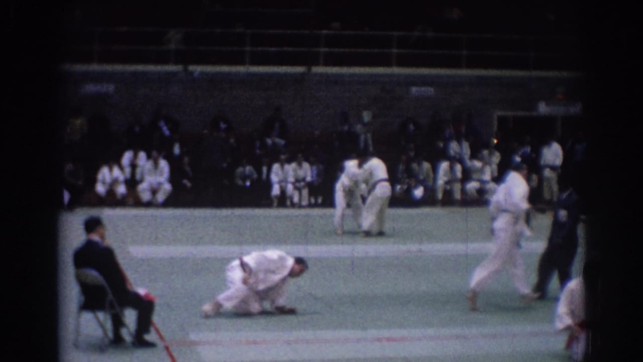 Indoor 1967 Los Angeles judo scene with one athlete on the mat, another pair standing, and spectators along the far side.