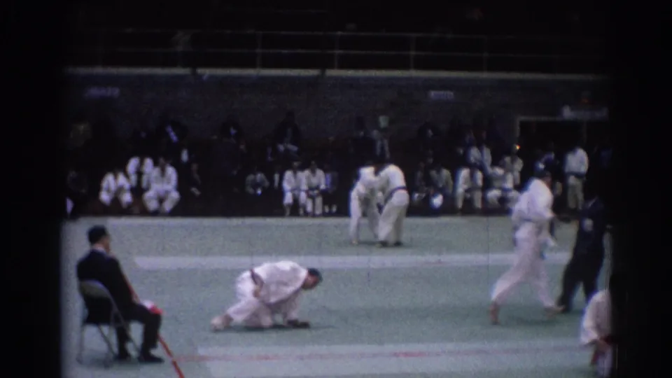 Indoor 1967 Los Angeles judo scene with one athlete on the mat, another pair standing, and spectators along the far side.