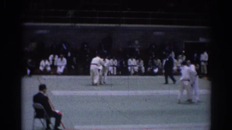 1967 Los Angeles gymnasium footage showing judo competitors on a green mat, an official seated nearby, and spectators behind them.