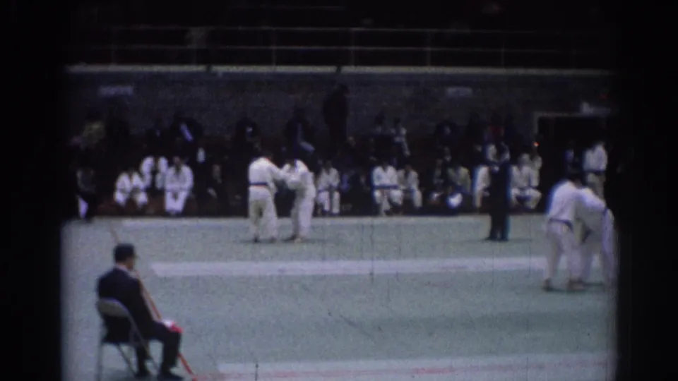 Late-clip frame from a 1967 Los Angeles judo competition with athletes standing on the mat and a dark-clothed official nearby.