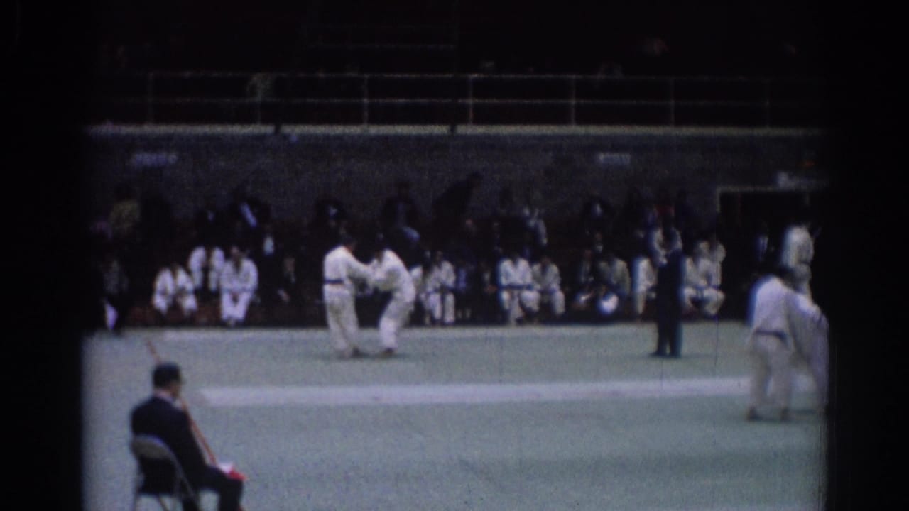 Two judo competitors in white uniforms on a green mat with spectators behind them.