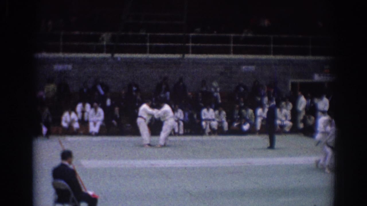 1967 Los Angeles frame of two judo competitors in white gi uniforms during a match, with spectators seated along the back wall.