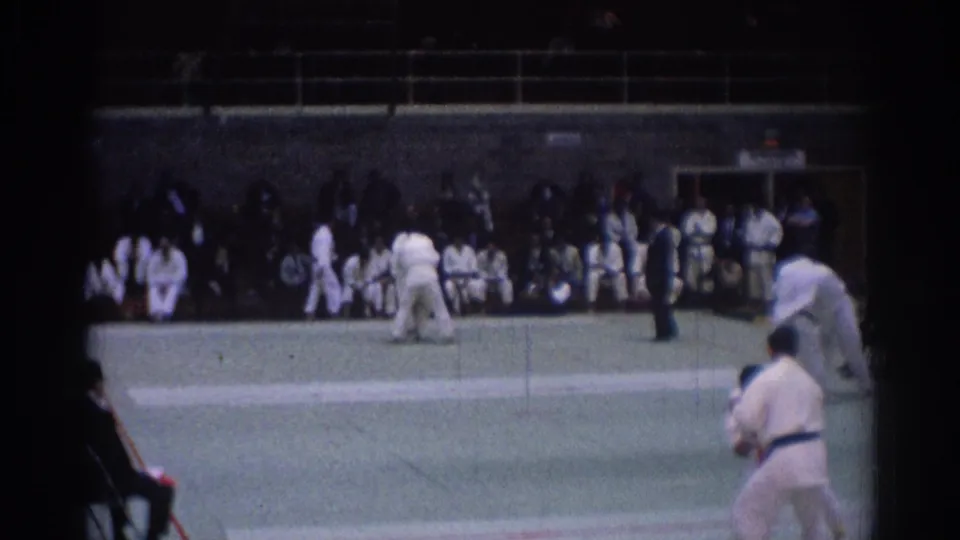 1967 Los Angeles judo competition frame showing competitors in action on the green mat, with spectators and officials around the floor.