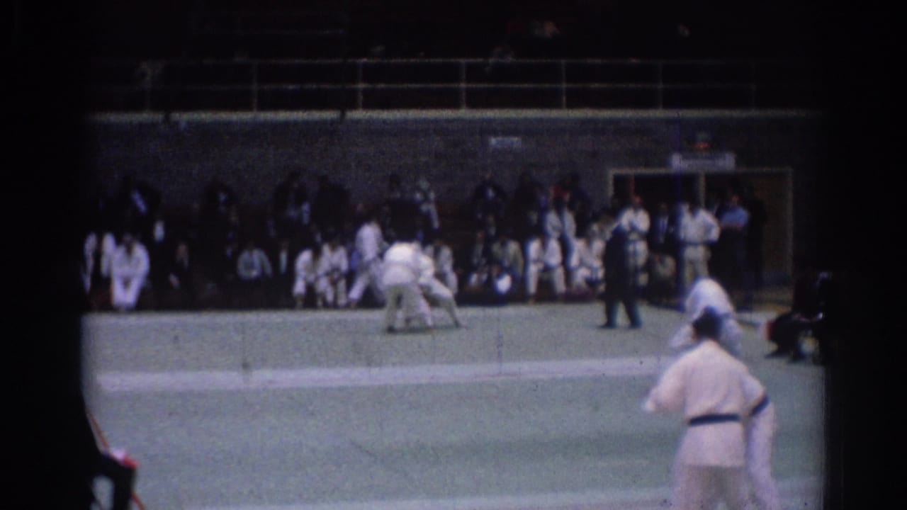 Two judo athletes grapple on a green mat while a referee and seated audience watch.
