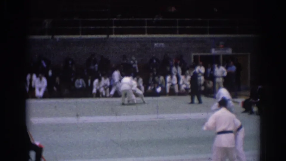 1967 Los Angeles 8mm frame showing two judo athletes in white gi grappling on an indoor mat before spectators and a referee.