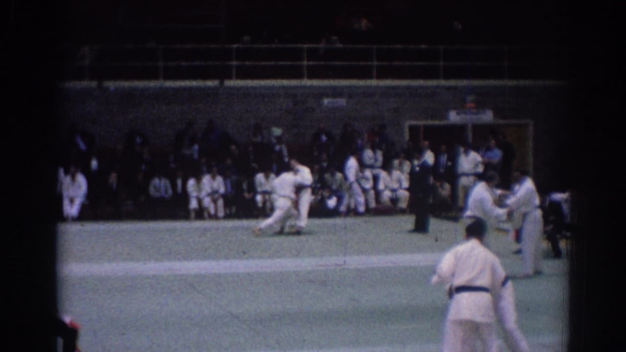 Indoor 1967 Los Angeles judo scene with one athlete low on the mat, another standing at right, and spectators watching from the back.