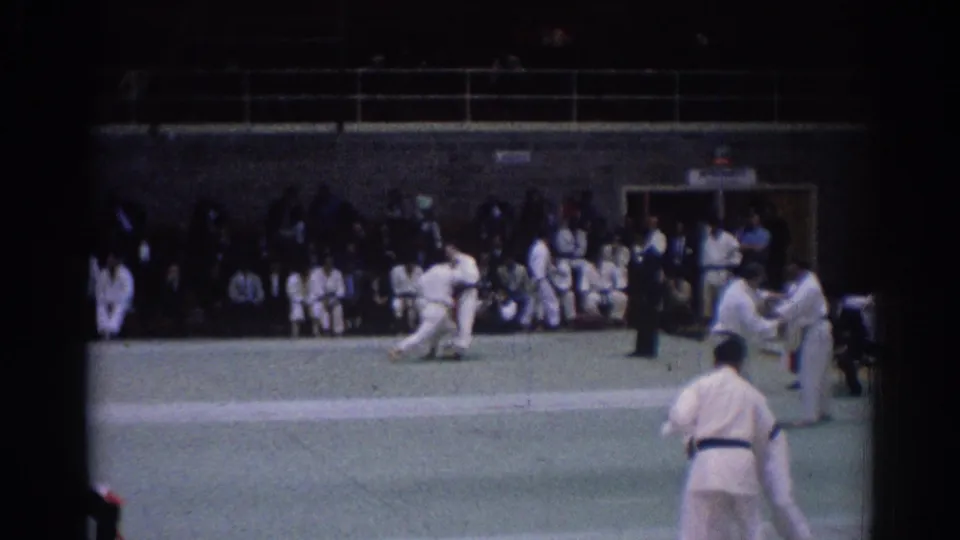 Indoor 1967 Los Angeles judo scene with one athlete low on the mat, another standing at right, and spectators watching from the back.