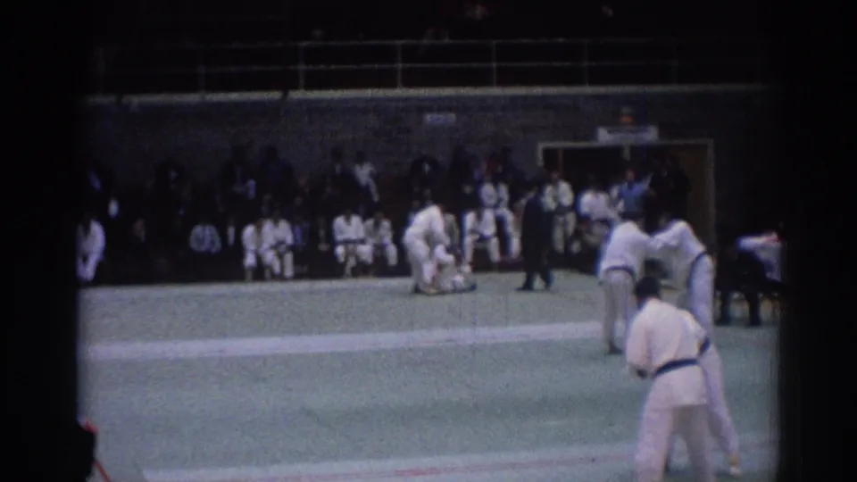 1967 Los Angeles indoor judo footage showing athletes in white gi near the mat edge, with a referee and seated audience behind them.