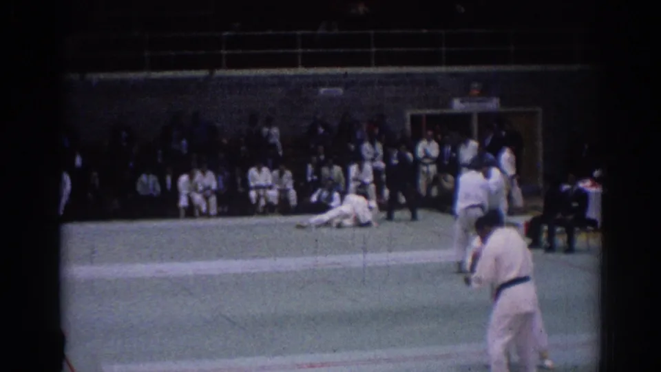 1967 Los Angeles 8mm frame of an indoor judo bout, with one athlete down on the mat and others standing near the referee.