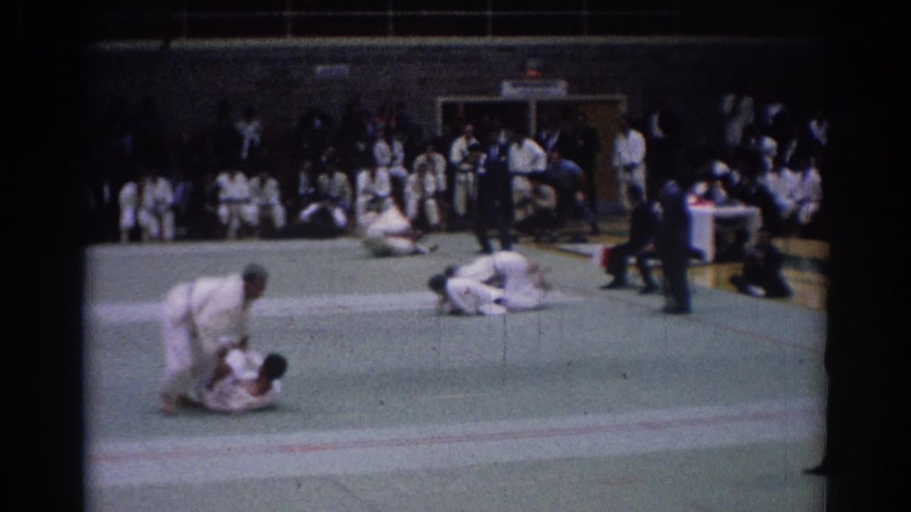 1967 Los Angeles home-movie frame showing gi-clad competitors grappling on a gym mat with spectators in the background.