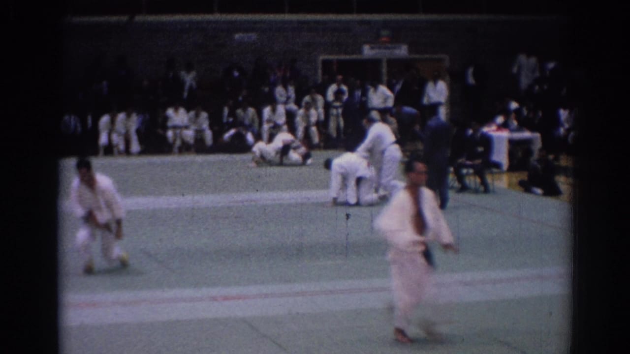 Competitors continue grappling while another gi-clad person crosses in the foreground.