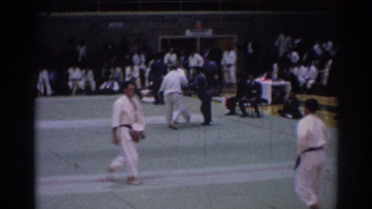 1967 Los Angeles gym tournament frame showing a gi-clad man crossing the mat near officials and waiting competitors.