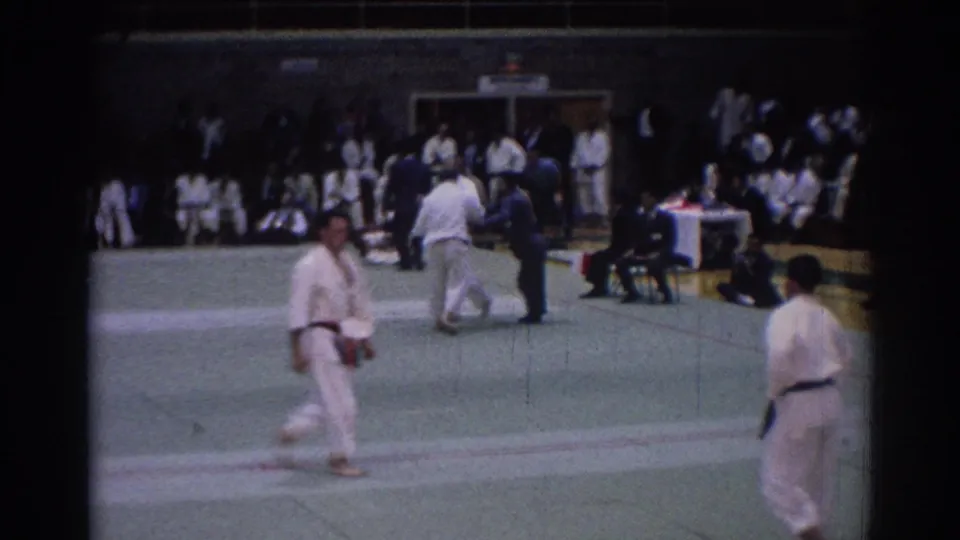 1967 Los Angeles gym tournament frame showing a gi-clad man crossing the mat near officials and waiting competitors.