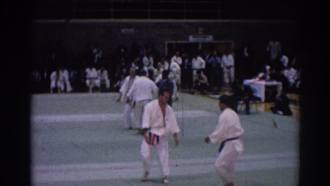 1967 Los Angeles home-movie frame showing martial arts competitors standing near the mat edge during a gym tournament.