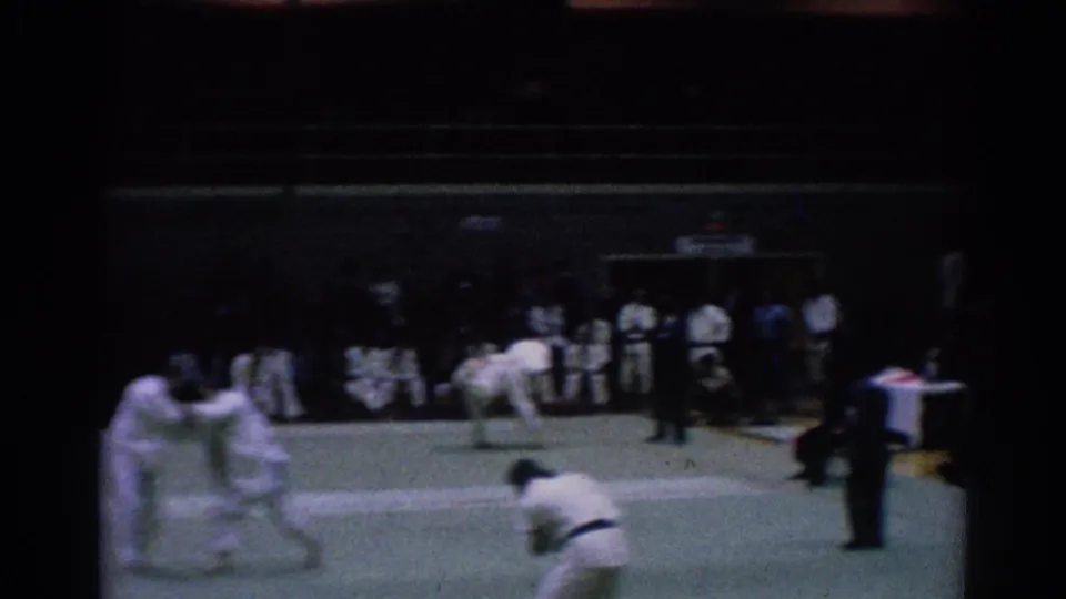 1967 Los Angeles 8mm frame of martial artists in white gi competing on a green mat with a referee and spectators nearby.