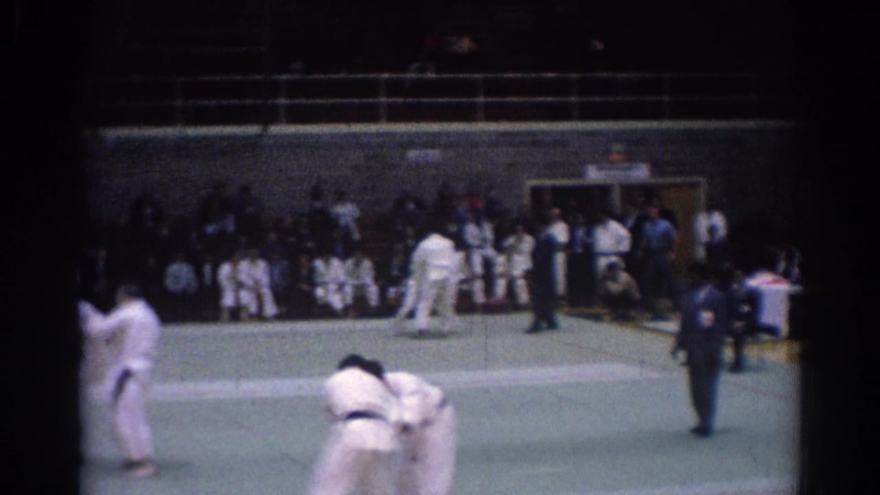 1967 Los Angeles home-movie frame showing a martial arts bout on a green mat with judges, spectators, and other participants visible.