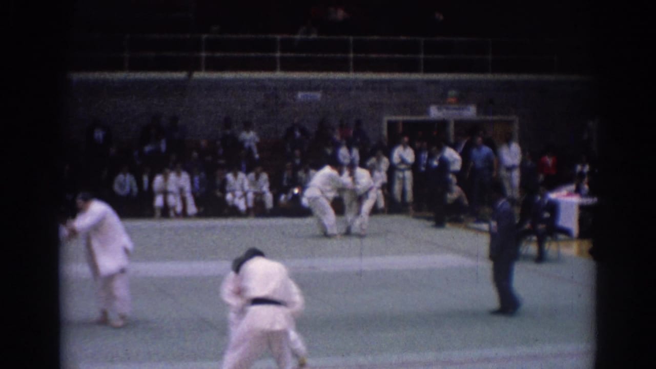 1967 Los Angeles home-movie frame of martial artists grappling on an indoor green mat with officials and spectators in view.