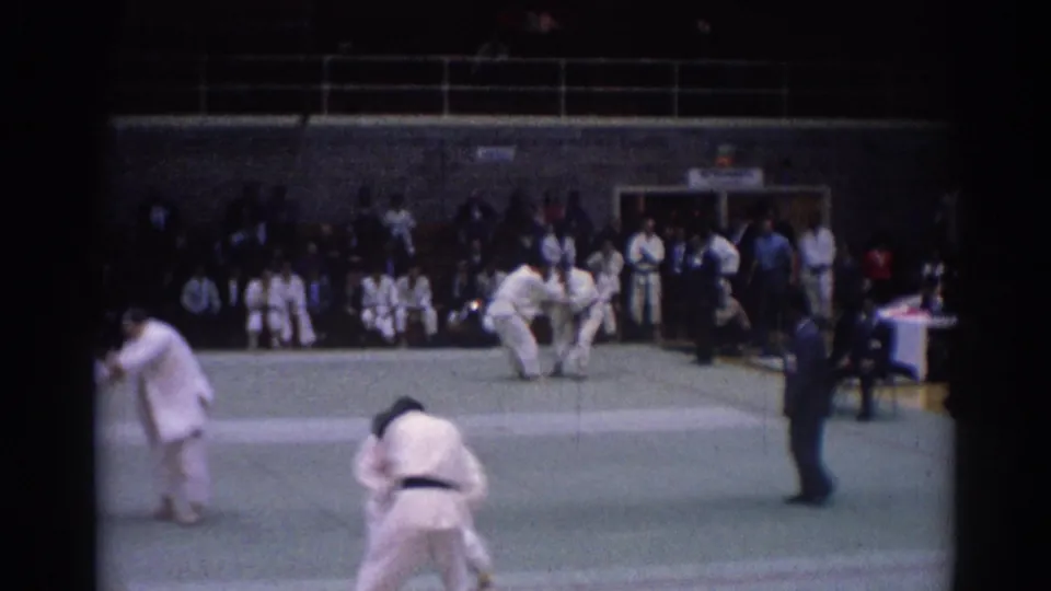 1967 Los Angeles home-movie frame of martial artists grappling on an indoor green mat with officials and spectators in view.