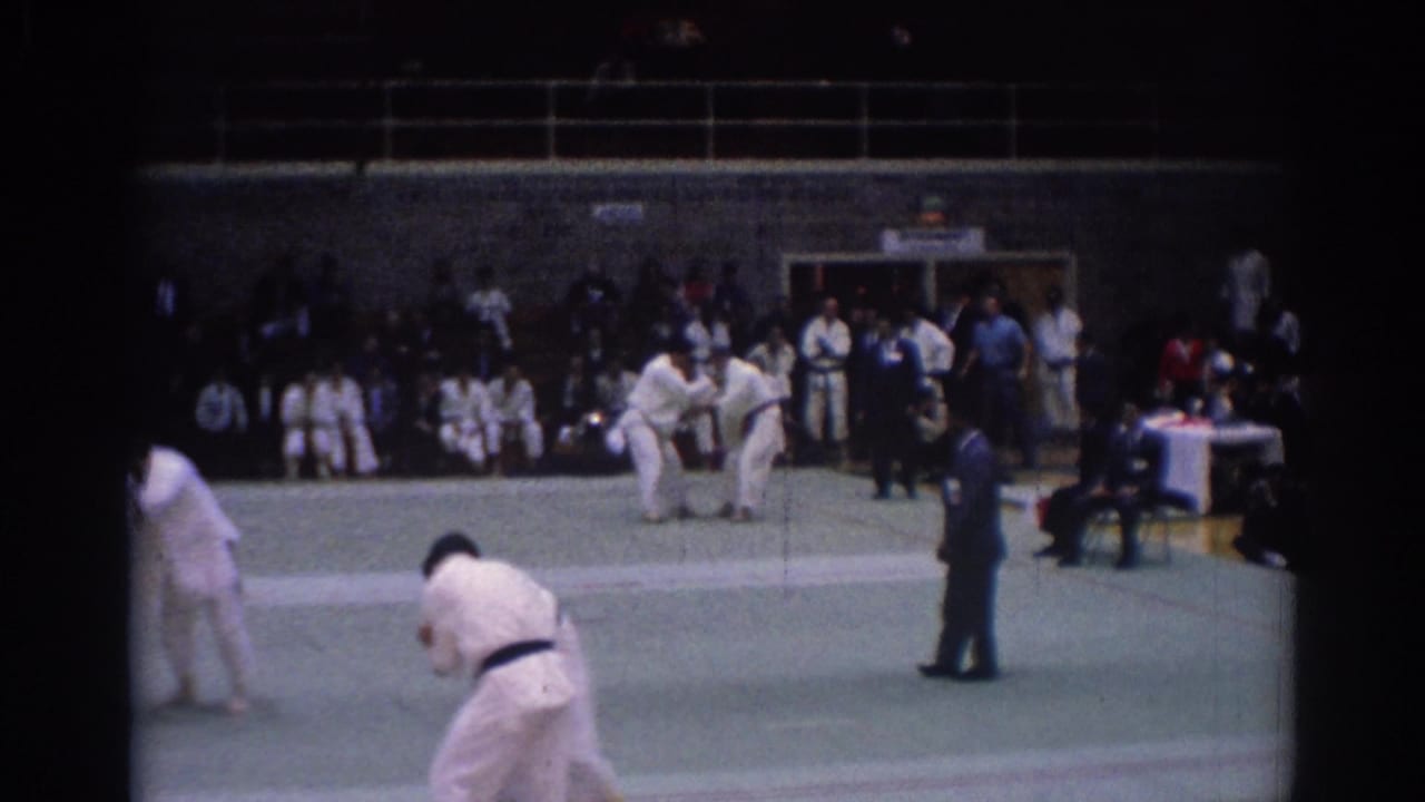 Men in white gi spar on a green mat while a referee and spectators watch.