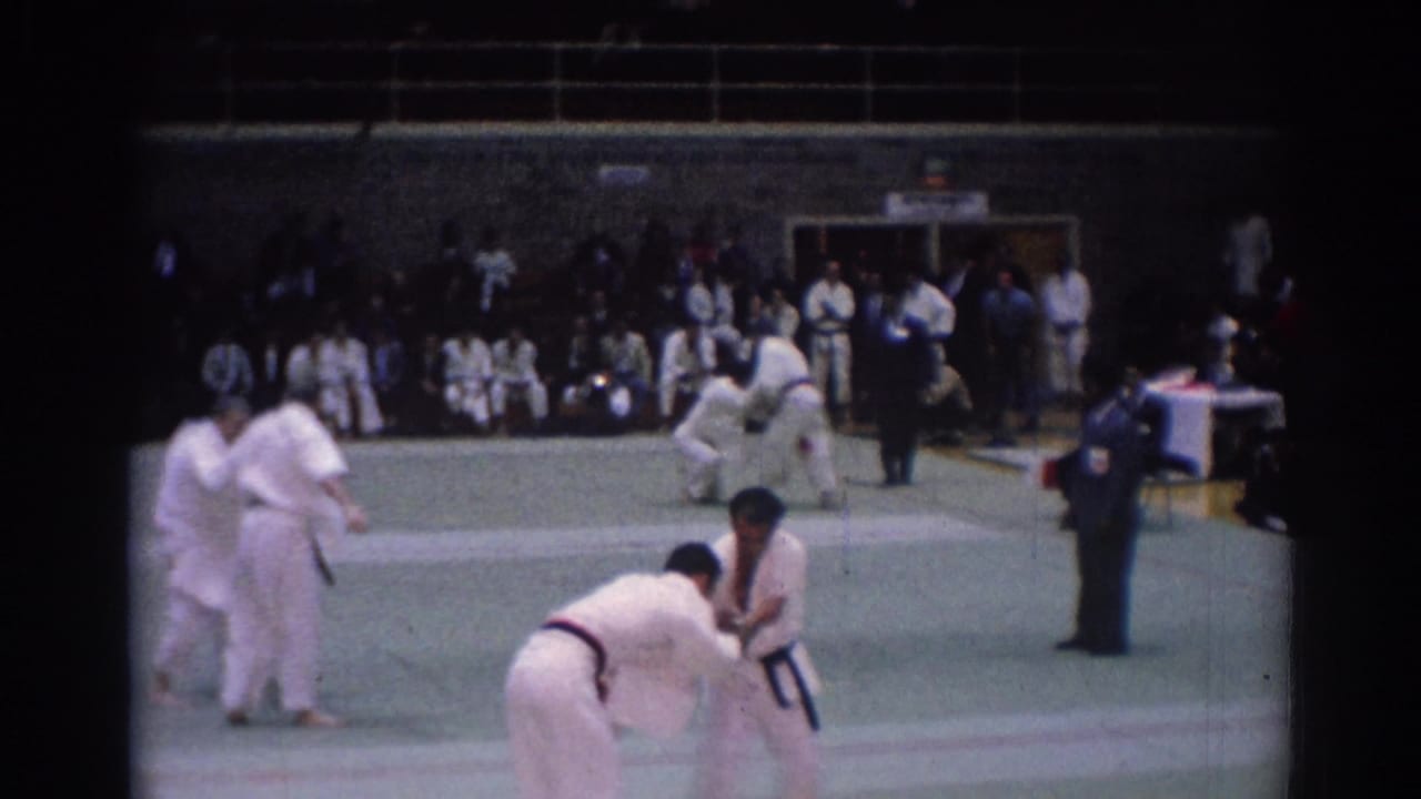 1967 Los Angeles 8mm footage frame with martial arts competitors in gi, sideline spectators, and multiple bouts on a green mat.