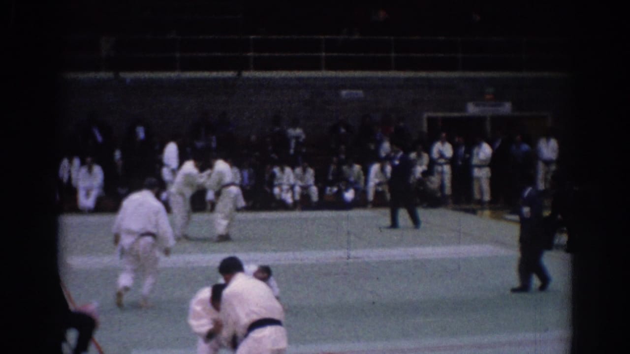 1967 Los Angeles martial arts competition frame showing several men in gi, a referee, and spectators inside a gym-like venue.