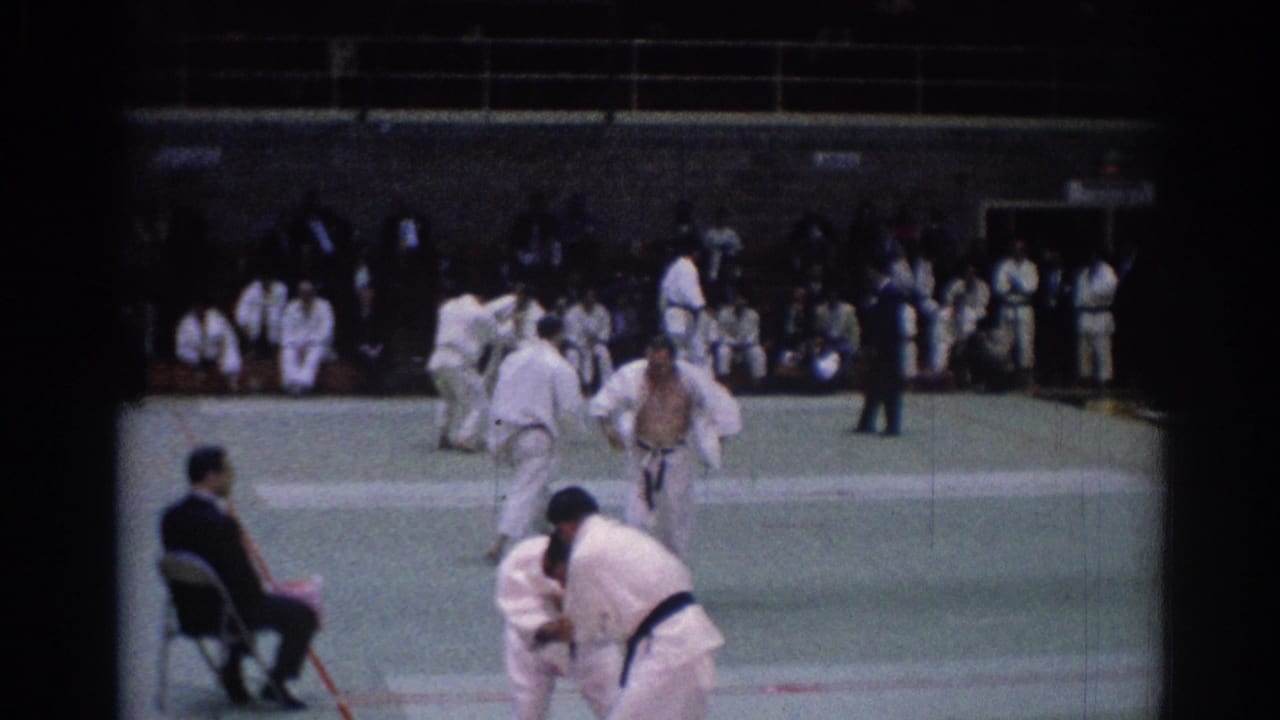 Judo competitors in white gi spar on a green mat while a seated judge and spectators watch.