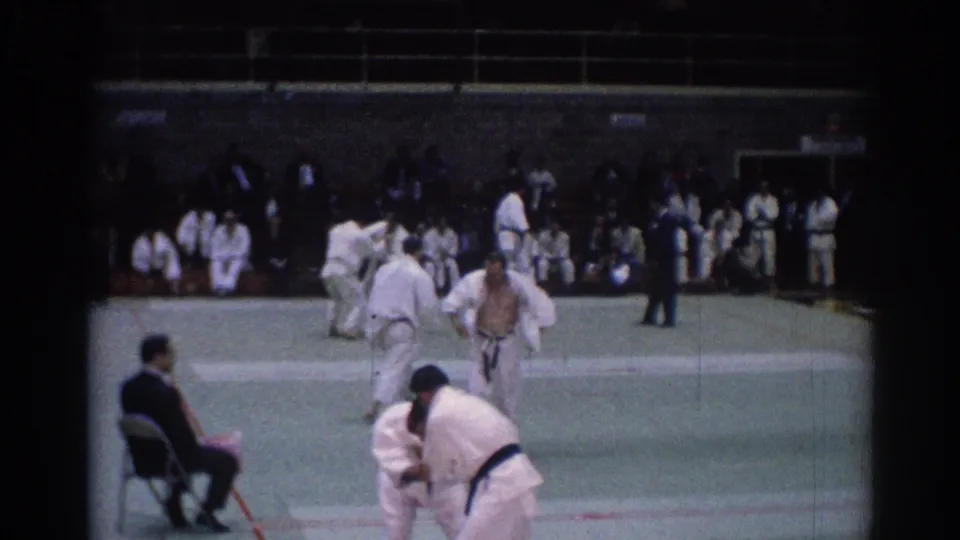 1967 Los Angeles 8mm frame showing judo competitors on an indoor mat with a judge seated nearby and spectators along the back wall.
