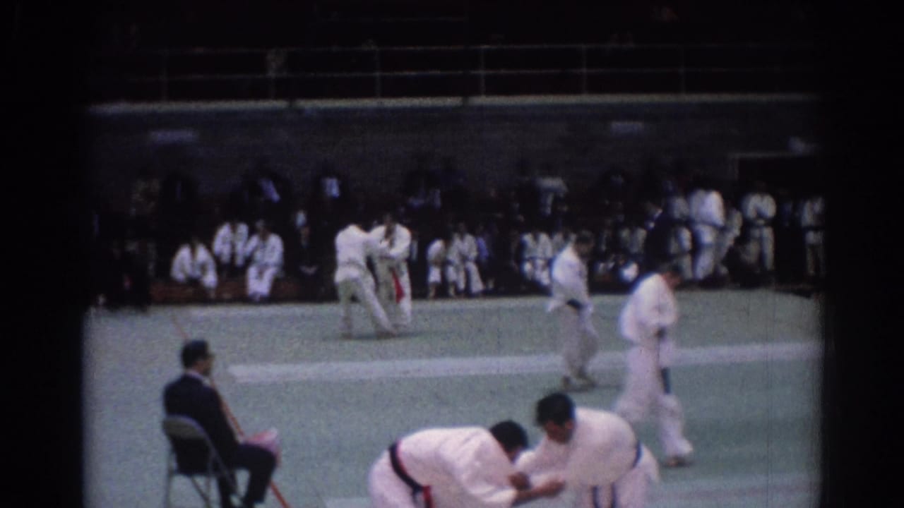 A 1967 Los Angeles judo match continues on an indoor green mat, with athletes in white gi and a judge seated at the side.
