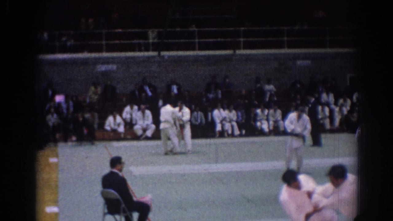 Indoor 8mm view of a 1967 Los Angeles judo tournament with players on the mat, a judge at left, and seated participants in the background.