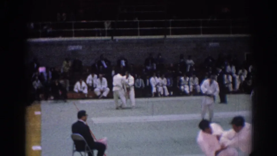 Indoor 8mm view of a 1967 Los Angeles judo tournament with players on the mat, a judge at left, and seated participants in the background.