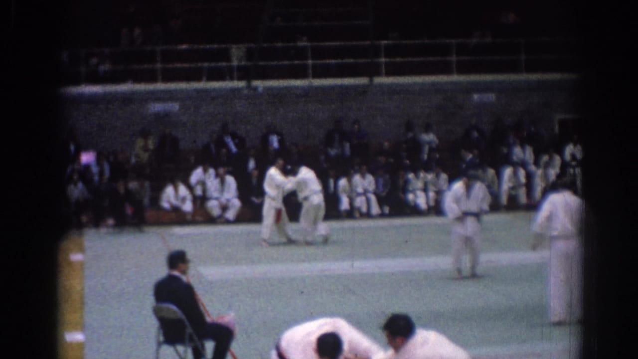 Late 1960s Los Angeles judo competition frame with grappling athletes, a seated judge, and rows of spectators in a large indoor space.