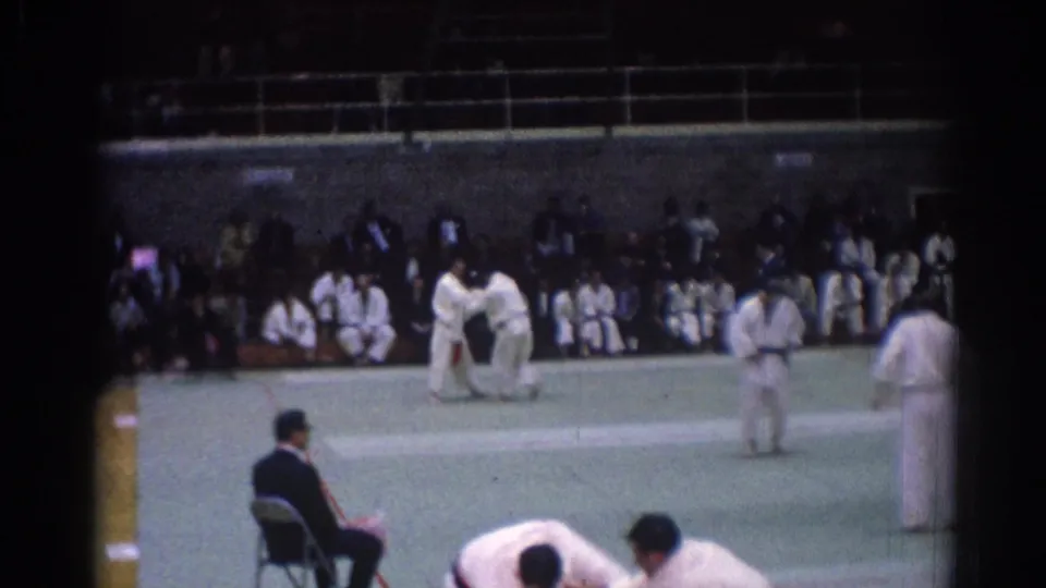 Late 1960s Los Angeles judo competition frame with grappling athletes, a seated judge, and rows of spectators in a large indoor space.