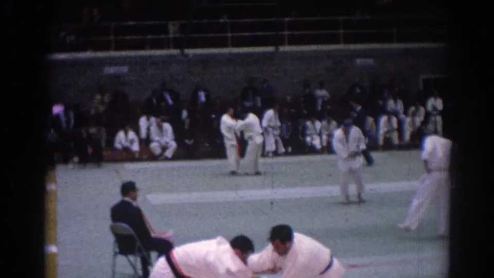 1967 Los Angeles home-movie frame showing judo competitors in white uniforms on a green mat as a judge and spectators watch.