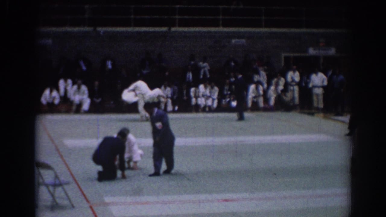 A 1967 Los Angeles judo competition frame shows an official signaling beside competitors on the mat with spectators in the background.