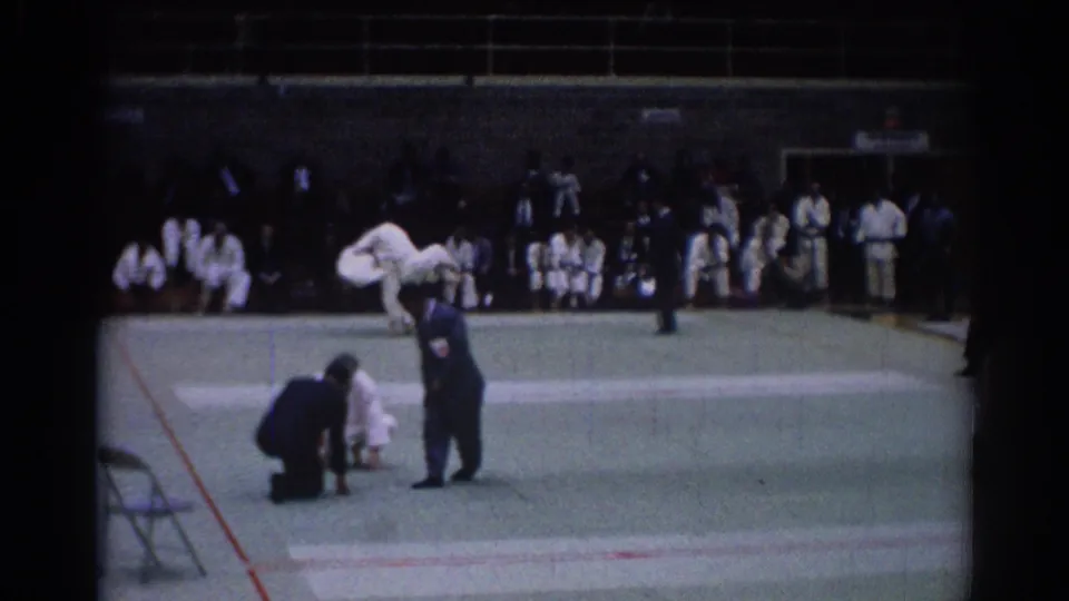 A 1967 Los Angeles judo competition frame shows an official signaling beside competitors on the mat with spectators in the background.