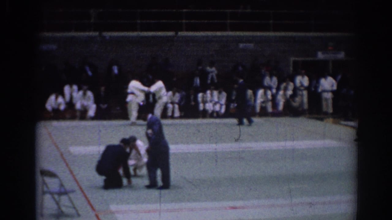 Two judo athletes grapple on a mat while a referee kneels nearby and spectators watch from the wall.