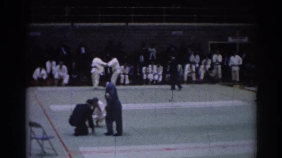 1967 Los Angeles home-movie frame of a judo bout with two athletes in white gi, a kneeling referee, and seated spectators in a gym.